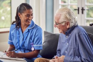 Health Care Assistant in blue uniform smiling and taking notes, sat next to an older male patient with white hair wearing a shirt and smiling.