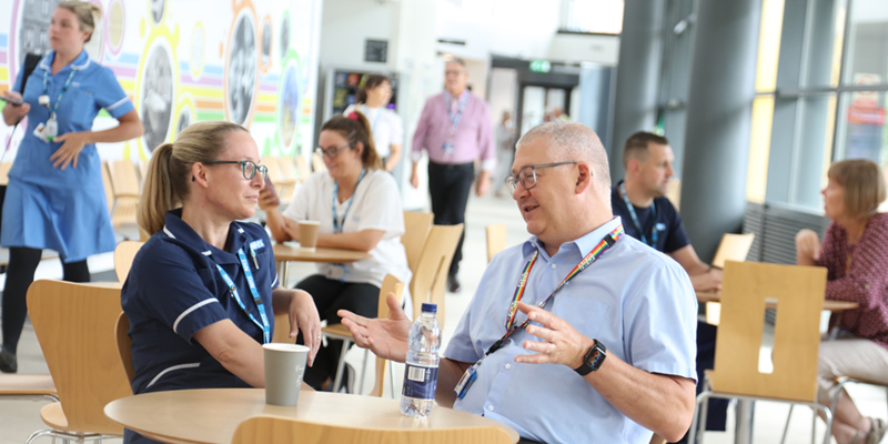 Colleagues talking and drinking coffee in a café area, with colleagues sitting and walking around them during a busy workday. 