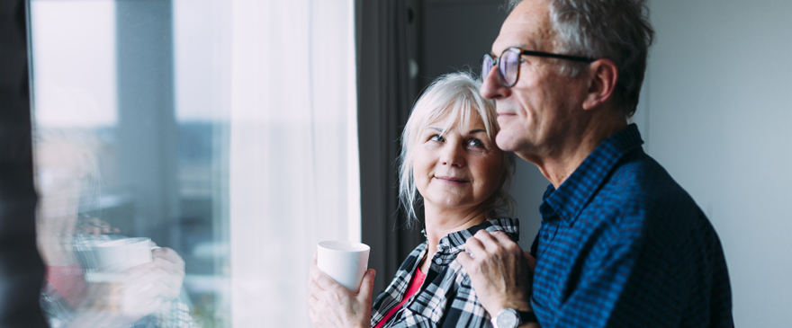 Man and woman standing next to each other by a window. The woman is holding a cup and looking at the man who is staring through the window.