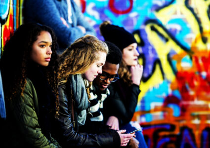 A group of teenagers are sitting in front of a wall covered in graffiti. They are wearing stylish clothes. A boy and girl are looking at a smartphone screen together.